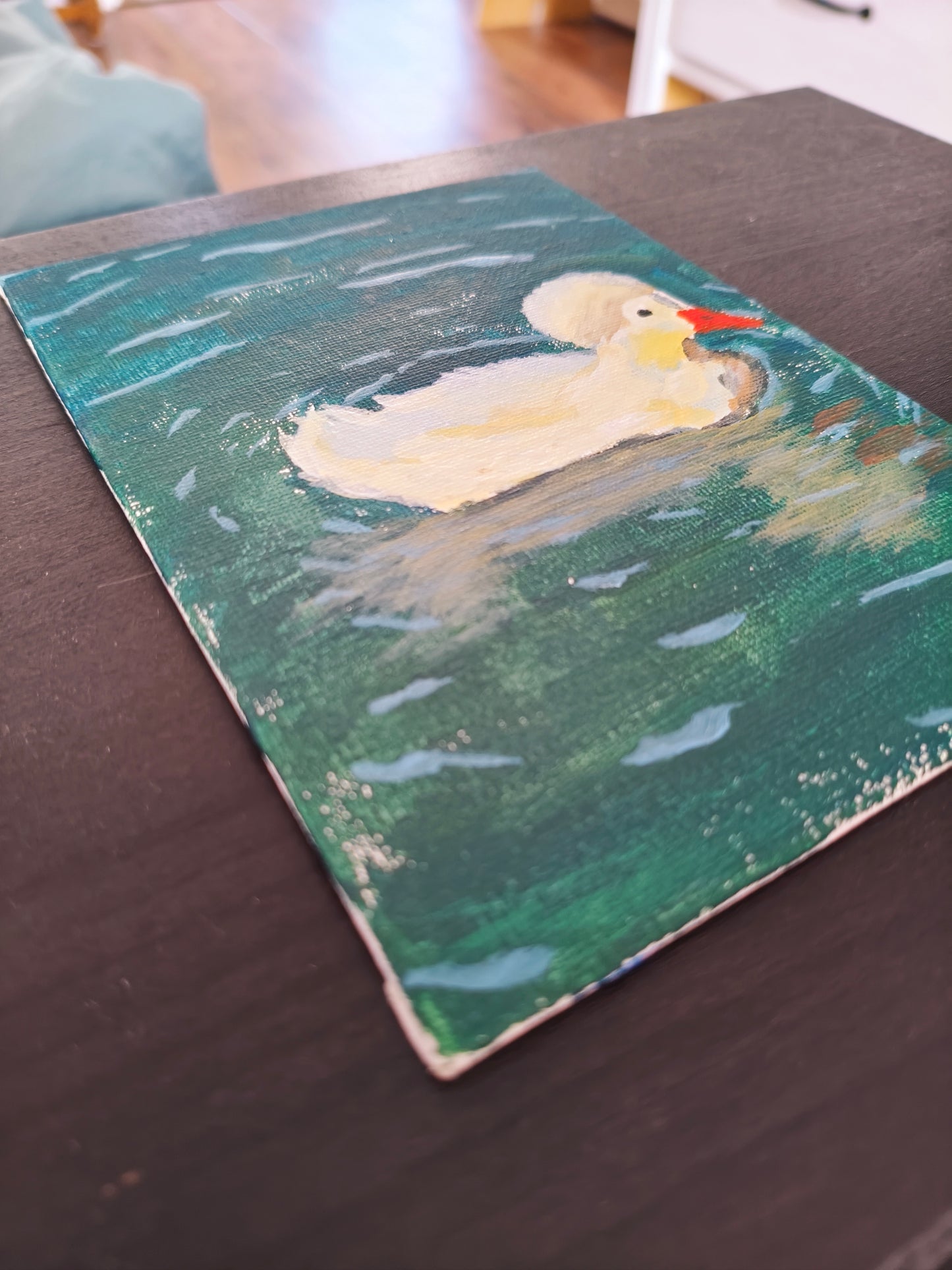 A rectangular painted canvas or print showing a white duck floating on green water, placed on a dark tabletop in an indoor setting.