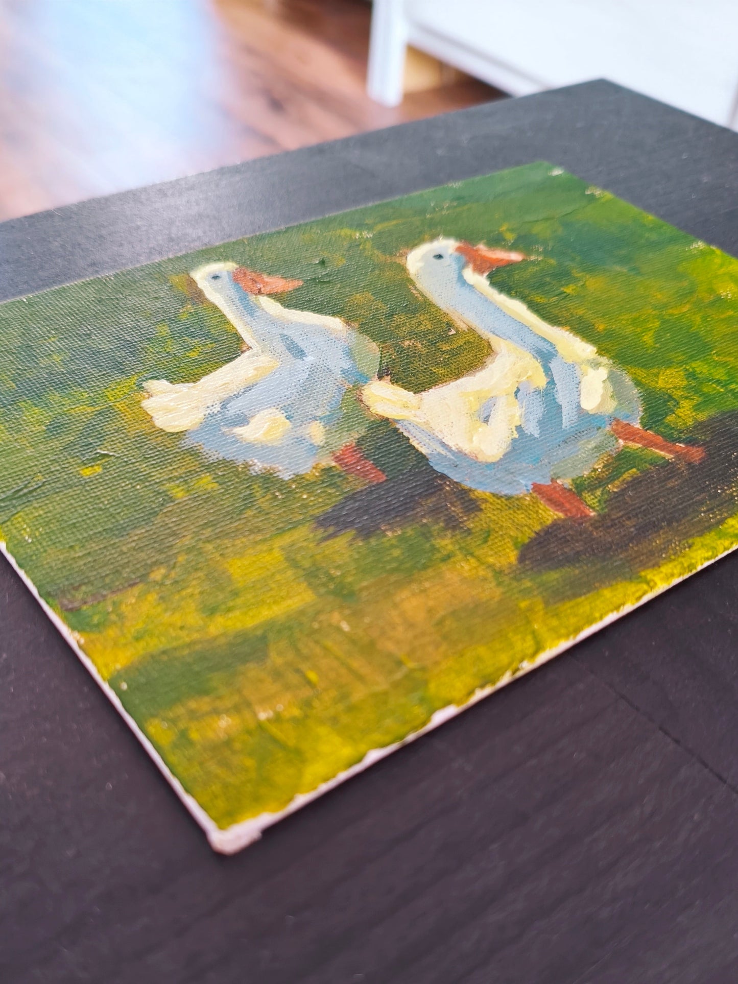 A small rectangular arcylic painting depicting two white geese with orange beaks on a textured green and yellow background, resting on a dark wooden table.