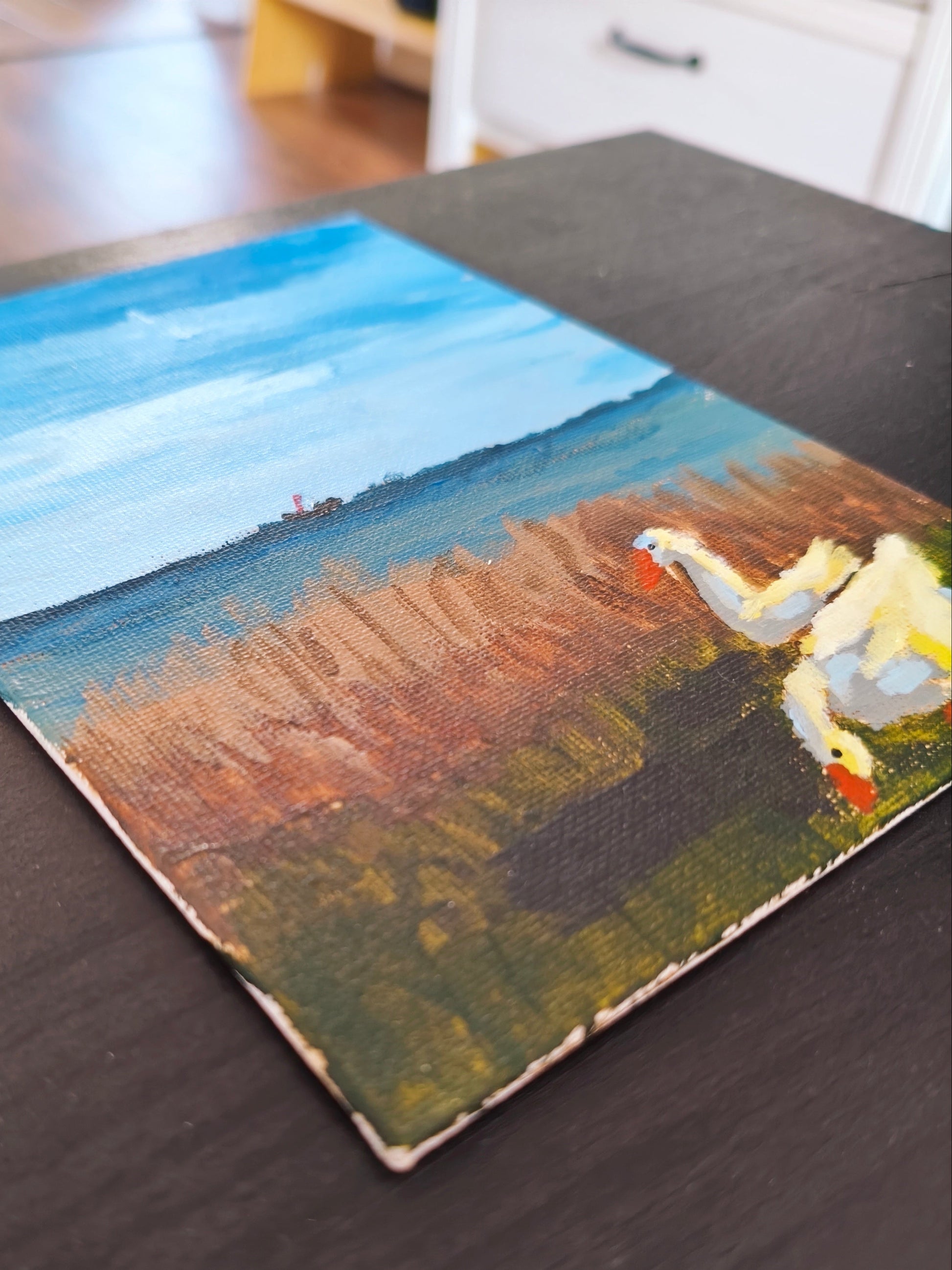 A small rectangular oil painting depicting a landscape with blue sky, brown and green fields, and white birds, resting on a dark wooden table.