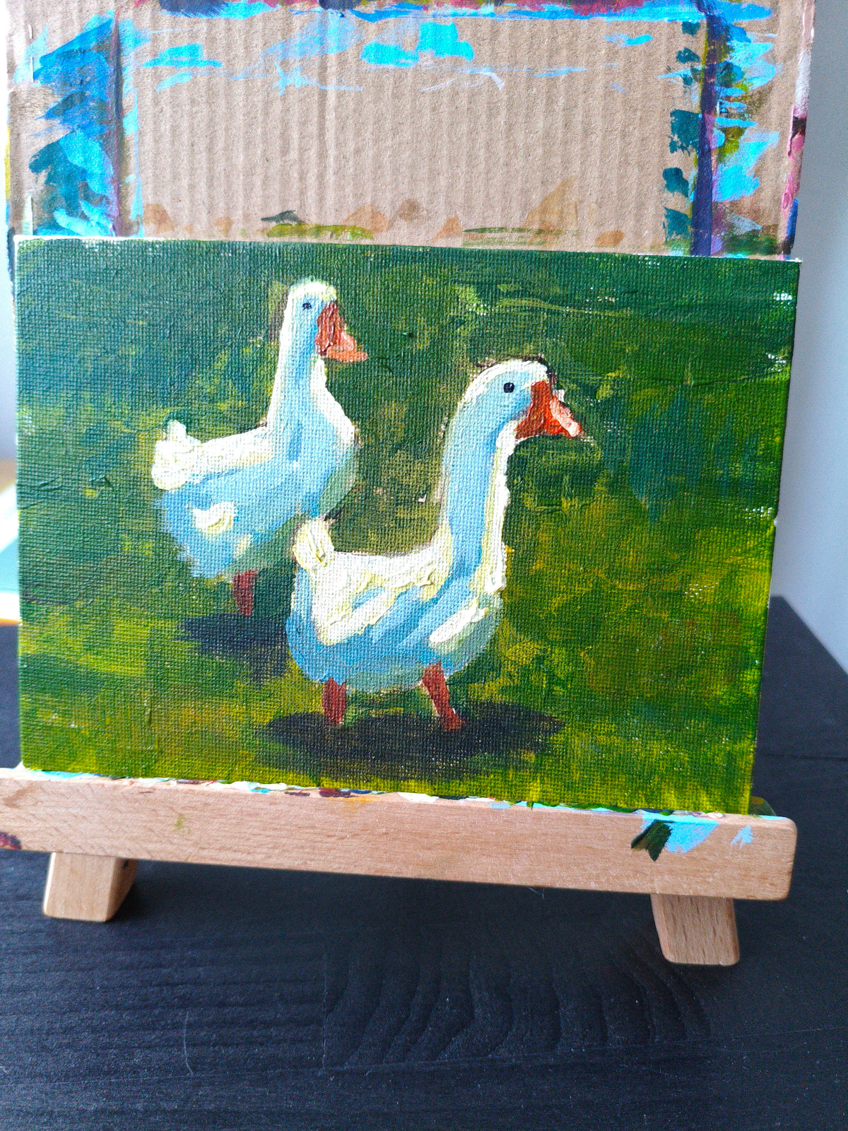 A small painted canvas showing two white geese on a green field, displayed on a wooden easel. The background behind the canvas shows a textured beige surface with blue paint marks.